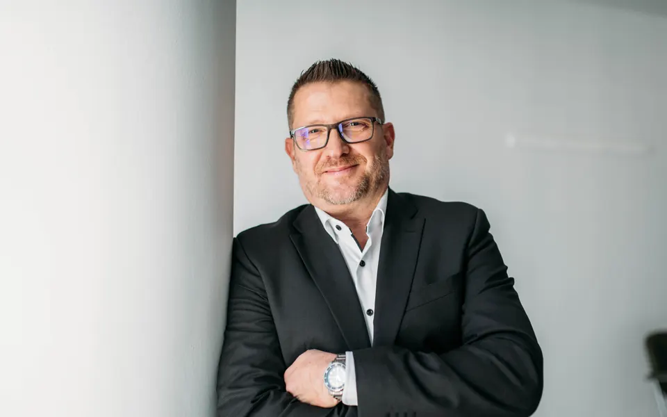 Andreas Pirker, Versicherungsmakler, lehnt mit verschränkten Armen an einer hellen Säule und lächelt in die Kamera